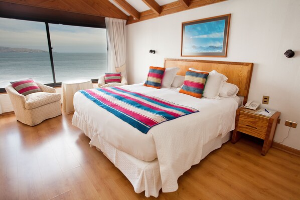 Une chambre avec un grand lit, une table de chevet en bois, une chaise et une vue sur la mer.