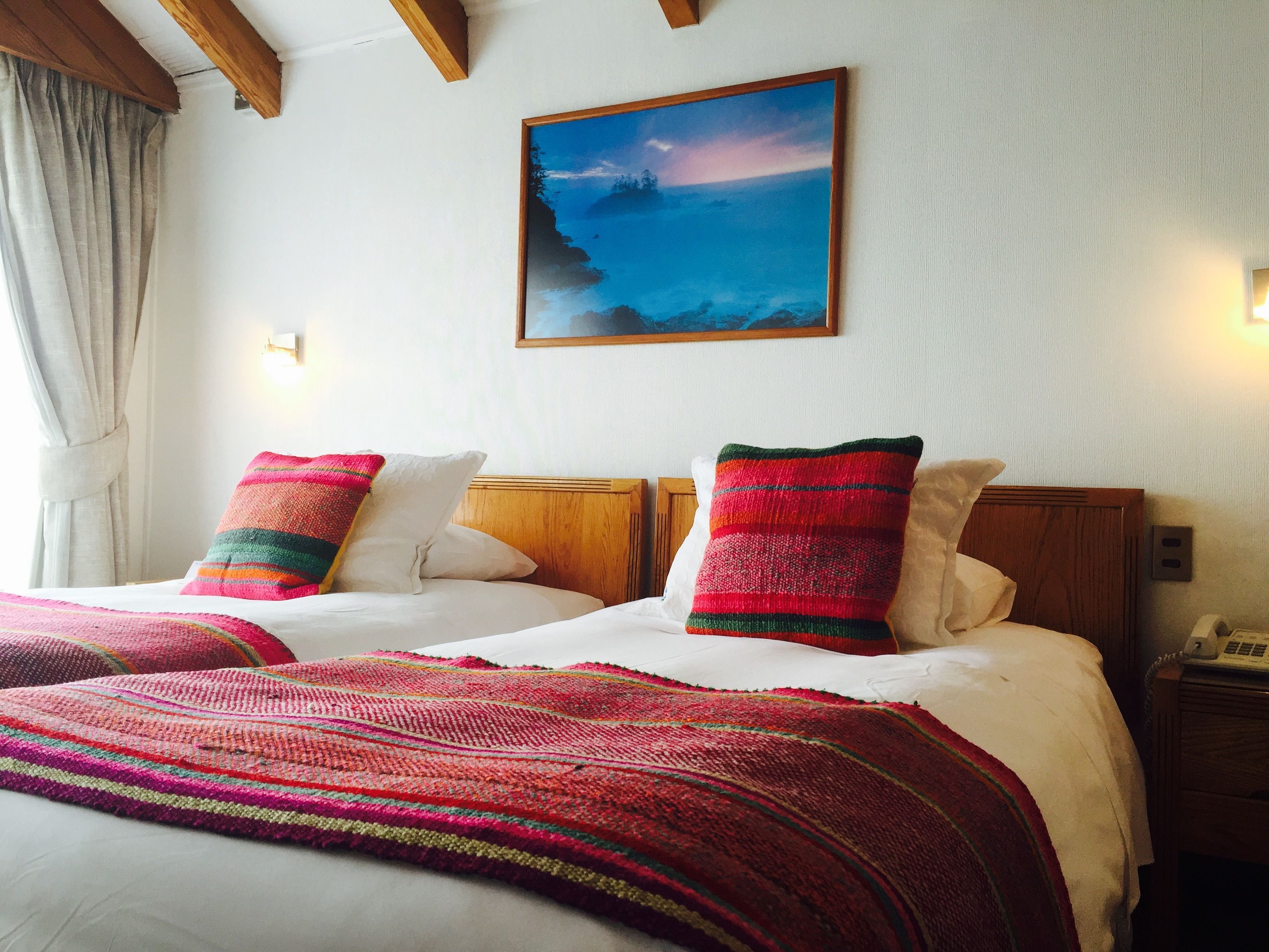 Uma cama de casal com duas almofadas coloridas, um quadro na parede acima e um telefone na mesa de cabeceira.