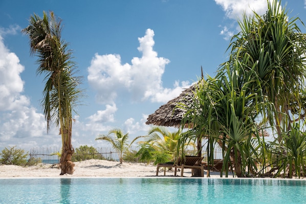 A pool area with a thatched-roof hut, palm trees, and a sandy beach.