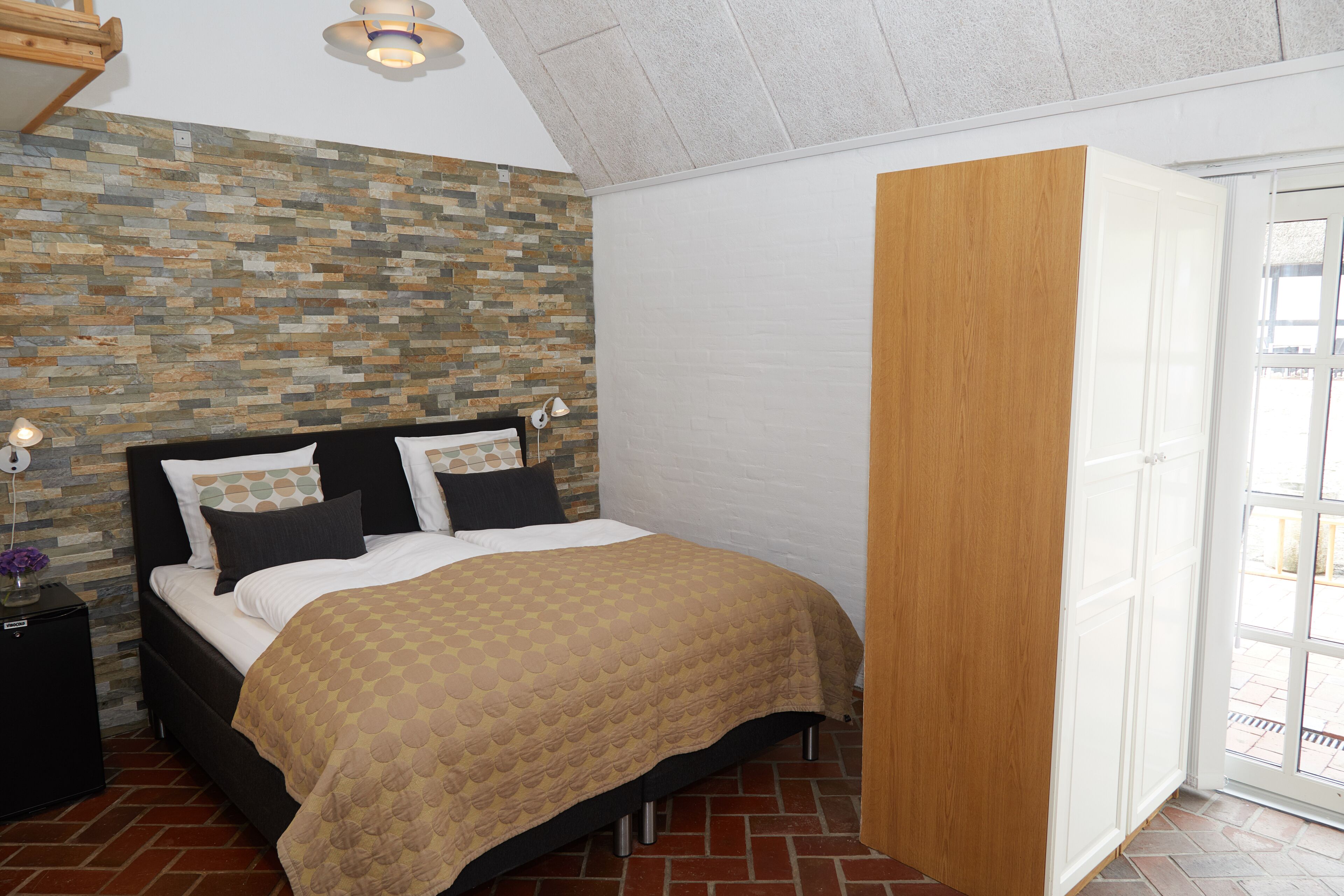 A bedroom with a bed, a nightstand, a wooden wardrobe, and a stone-patterned wall.