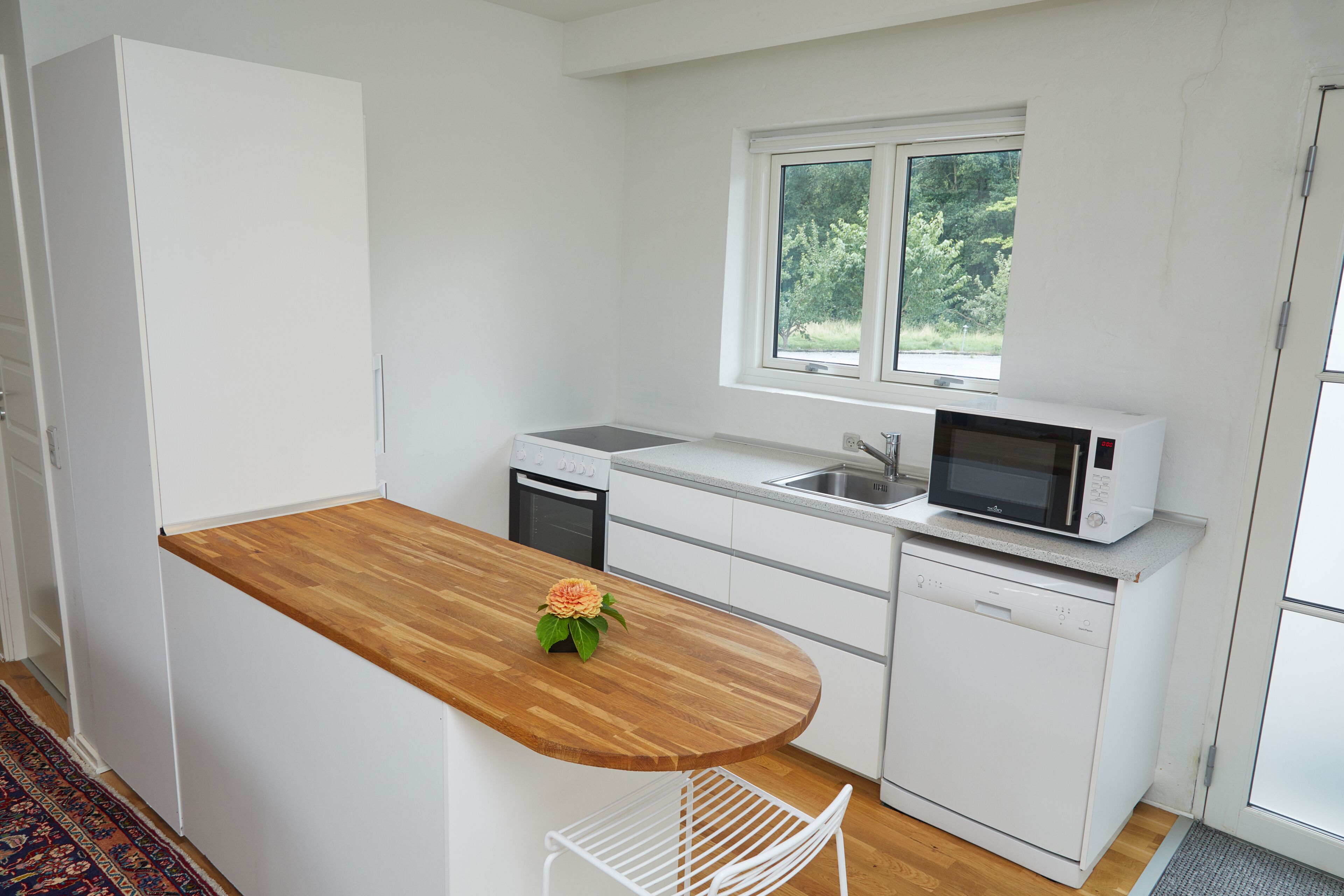 A compact kitchen with white cabinetry, a wooden countertop, a microwave, a dishwasher, and a window providing natural light.