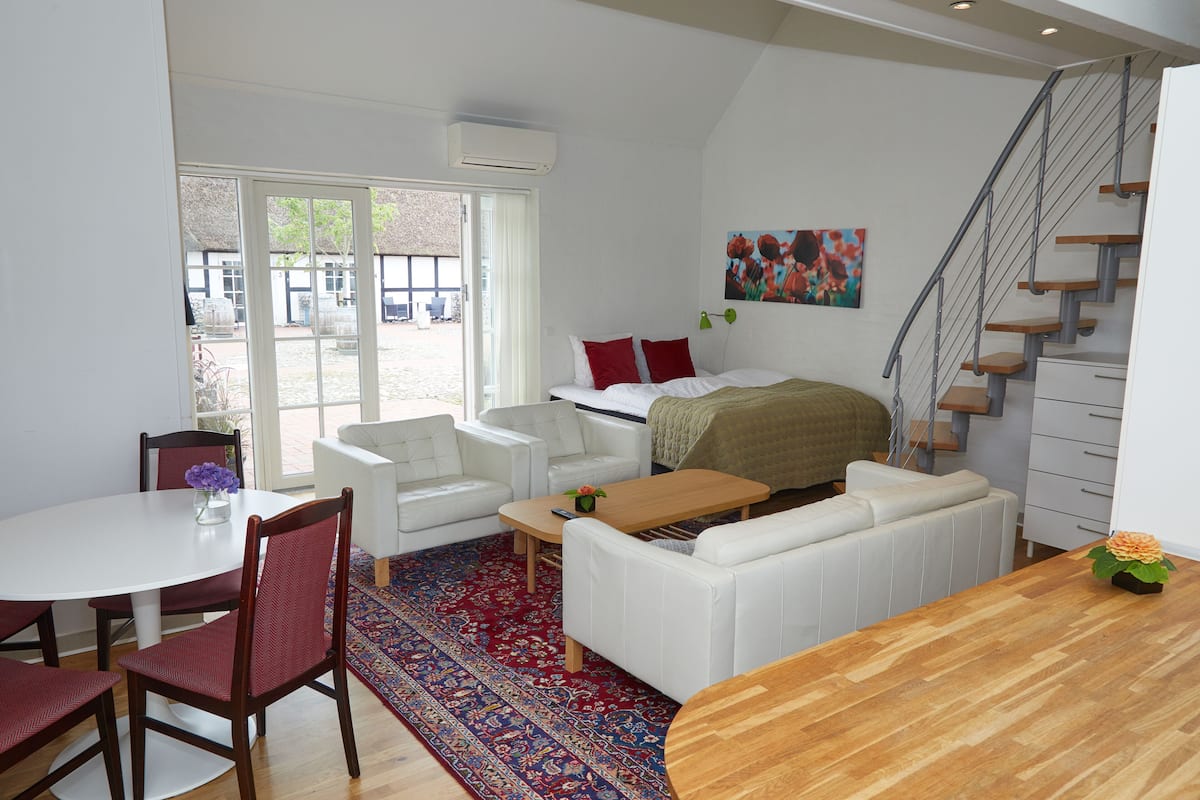 A modern, well-lit room with a dining area, a sofa set, a wooden table, and a staircase.