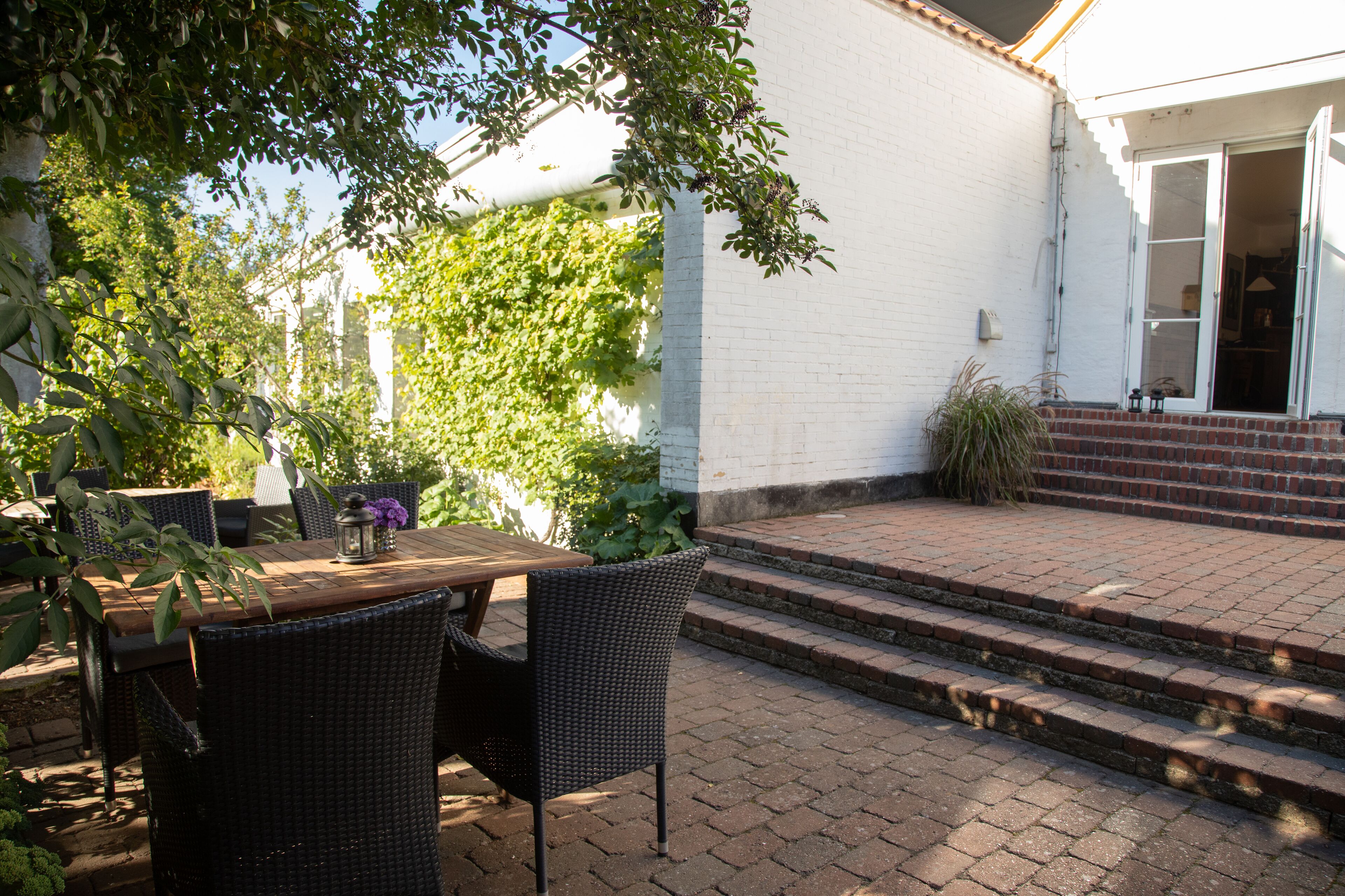 Eine Terrasse im Freien mit Tisch und Stühlen, ein Ziegelweg, der zu einer Tür führt, und ein weißes Gebäude im Hintergrund.
