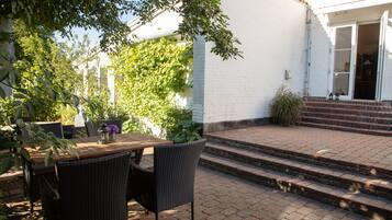 Eine Terrasse im Freien mit Tisch und Stühlen, ein Ziegelweg, der zu einer Tür führt, und ein weißes Gebäude im Hintergrund.