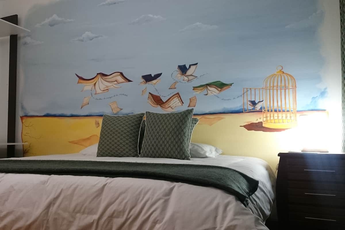 Une chambre à coucher comprenant un lit, des oreillers, une table de chevet et une fresque murale représentant des oiseaux et une cage.