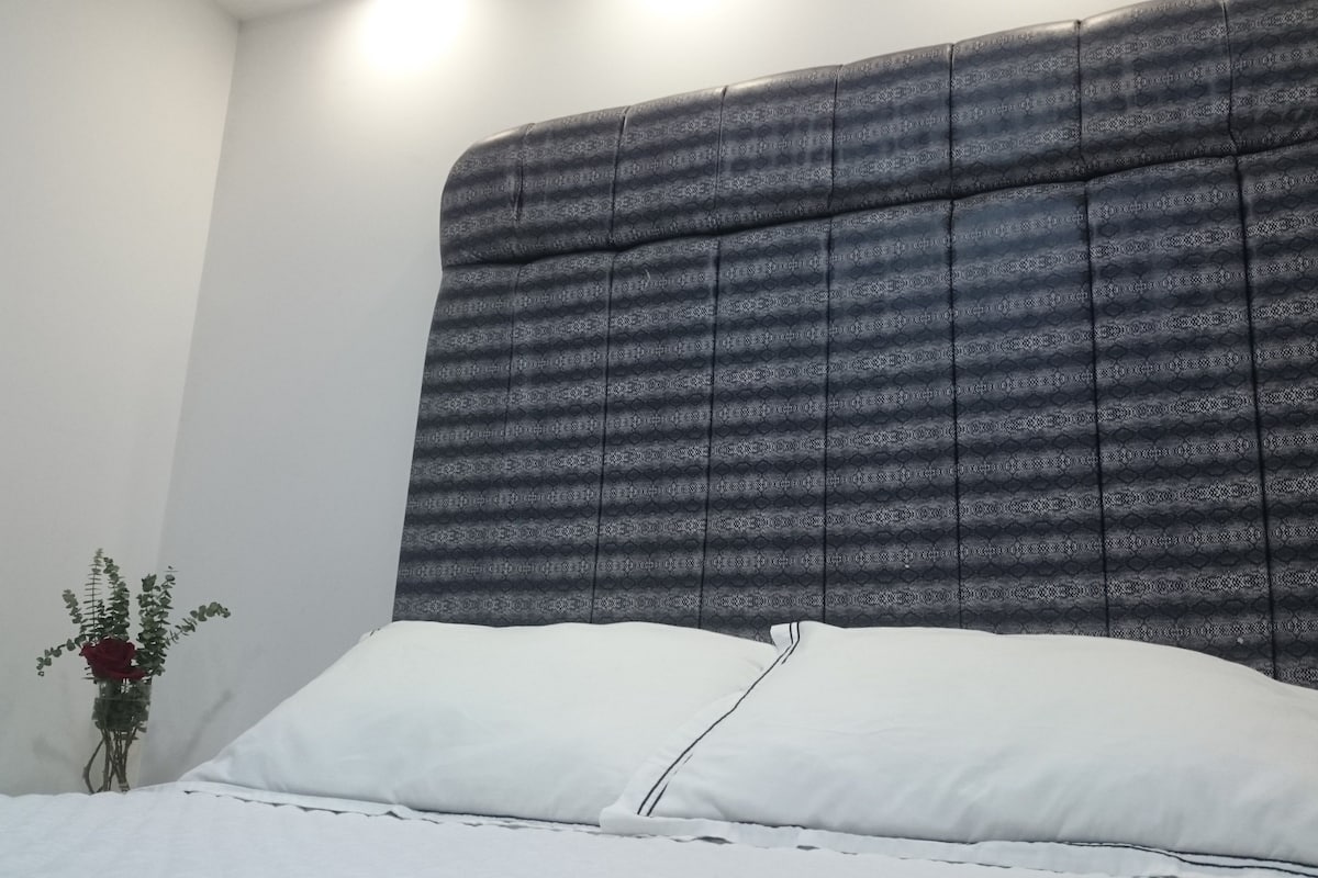 Un lit avec un matelas et des oreillers blancs, une tête de lit sombre à motifs et un petit vase avec une fleur rouge sur le côté.