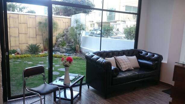 A living room with a leather sofa, a glass coffee table, and a chair. There is a view of a garden with a bamboo fence and a small pond. - Hotel + Arte (Quito)