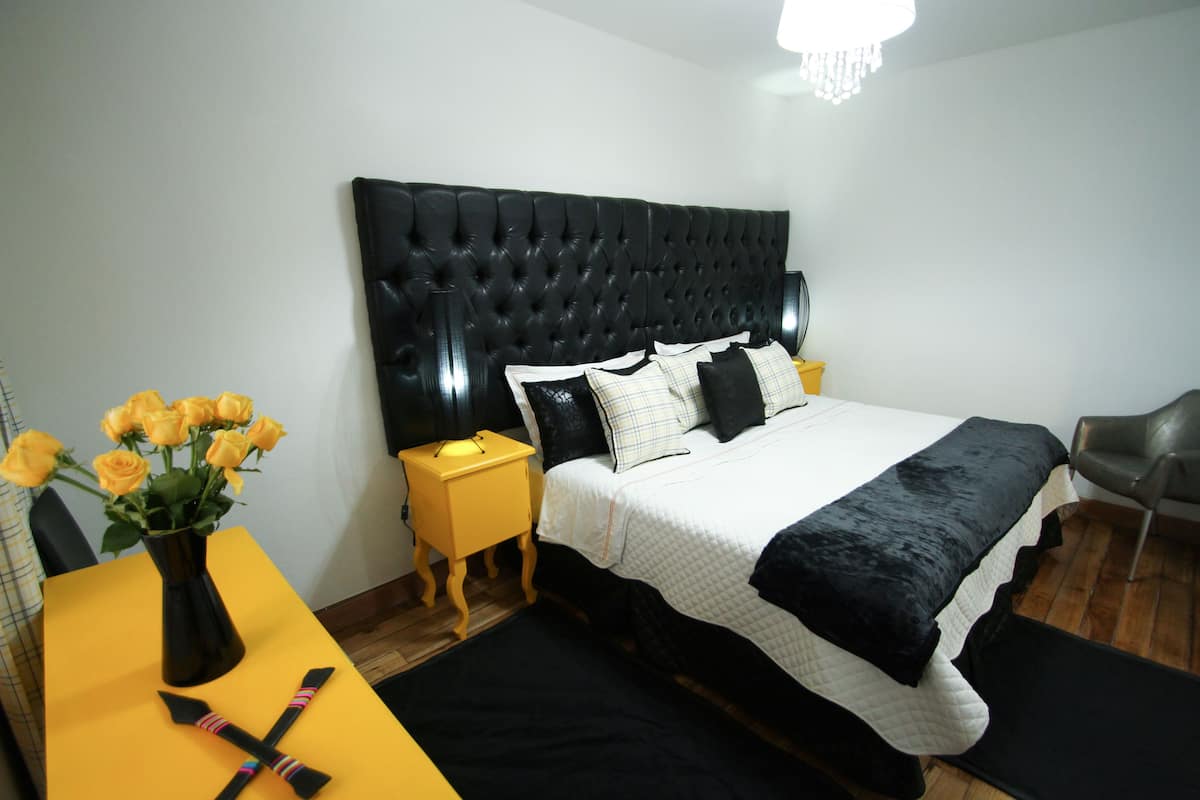 Une chambre à coucher avec une tête de lit capitonnée, un lit avec une literie noire et blanche, une table de chevet jaune sur laquelle est posé un vase de roses jaunes, et un tapis noir.