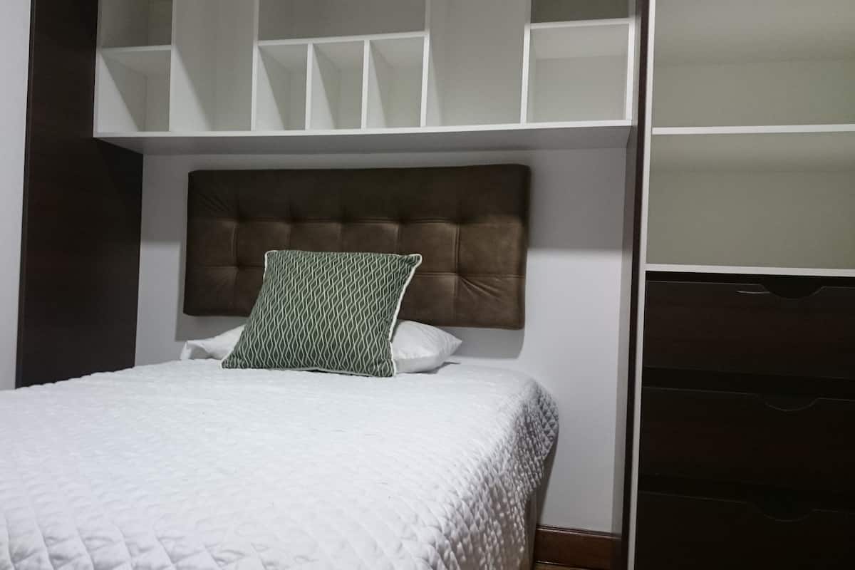 Un lit bien fait, recouvert d’une courtepointe blanche matelassée, agrémenté d’un coussin à motifs verts et doté d’une tête de lit capitonnée.