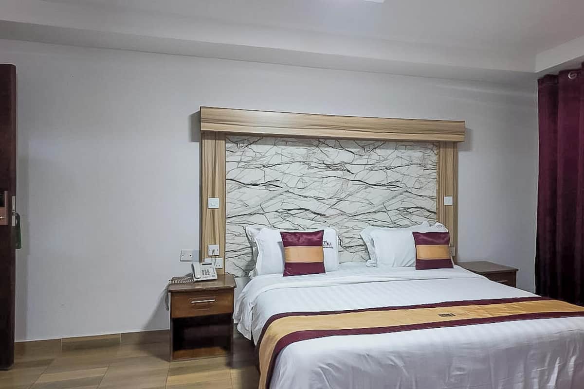 A hotel room with a marble-patterned headboard, a bed with white bedding and two decorative pillows, a bedside table with a phone, and a dark wooden wardrobe.