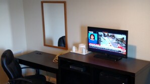 A hotel room with a desk, a television displaying news, a mirror, and a coffee cup.