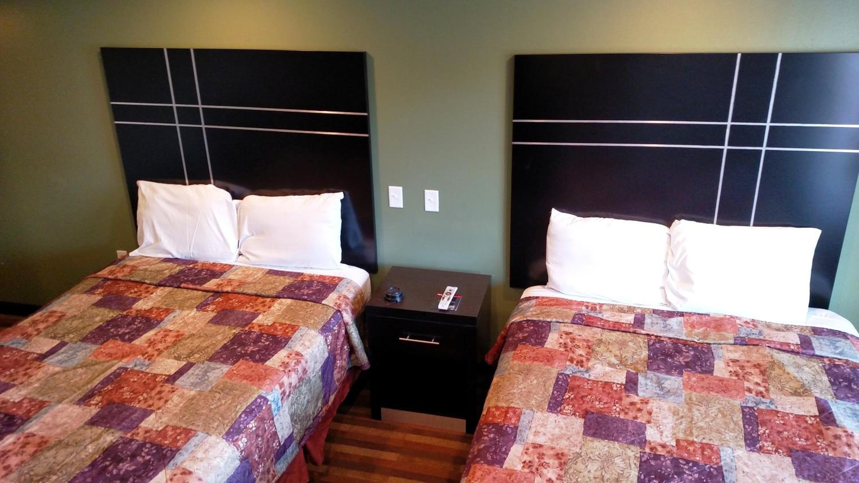 Two twin beds with matching headboards and white bedding, a small bedside table with a remote control, and a wall-mounted light switch.