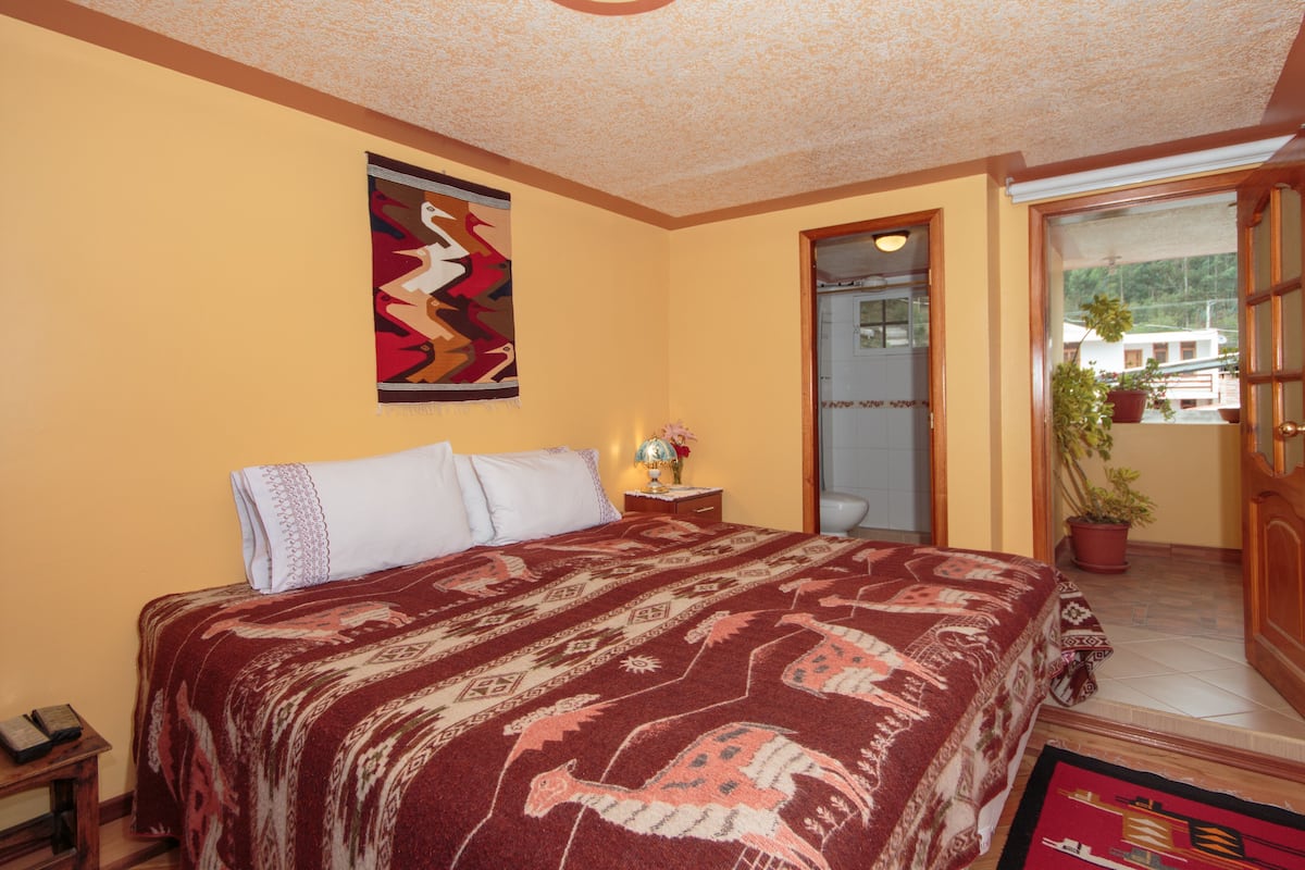A bedroom with a large bed, a nightstand, a wall hanging, and a door leading to a balcony with a potted plant.