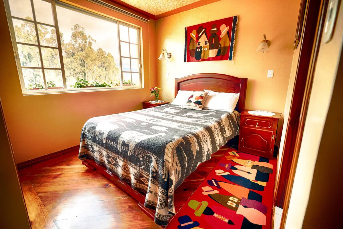A bedroom with a large bed, a window with a view of trees, a painting on the wall, and a colorful rug.