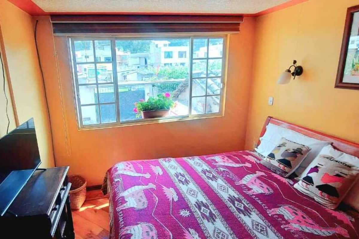 A bedroom with a bed, a desk, a window with a view, and a wall with a painting.