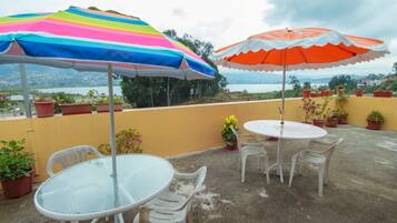 A patio with two round tables, white plastic chairs, and two colorful umbrellas. There are potted plants and a view of the landscape in the background.