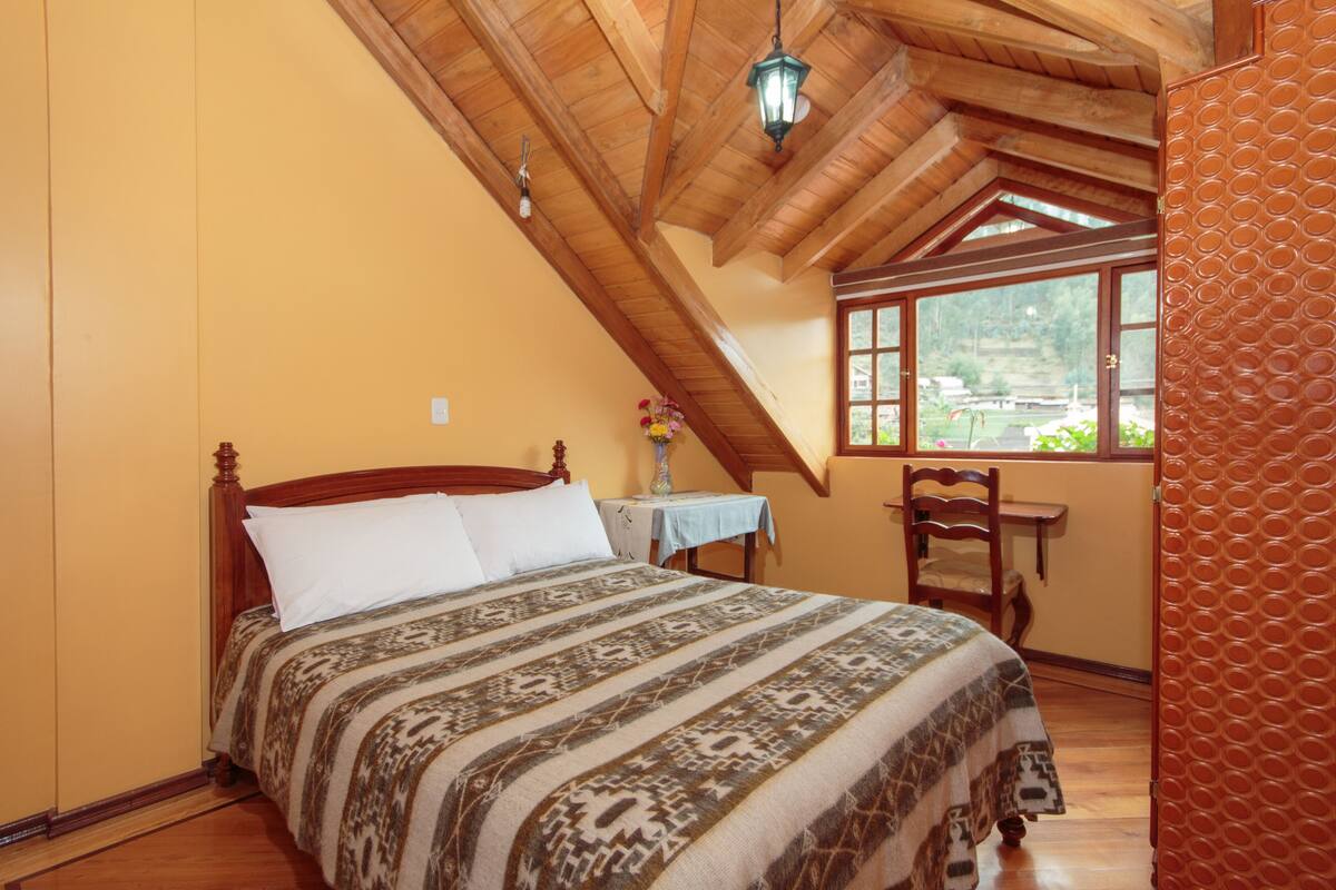 A bedroom with a wooden ceiling, a bed with patterned bedding, a small table with a vase, and a window with a view of greenery.
