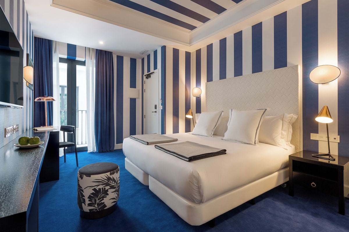 Habitación de hotel con una cama grande, mesitas de noche, un escritorio con silla y vistas al exterior a través de una ventana.