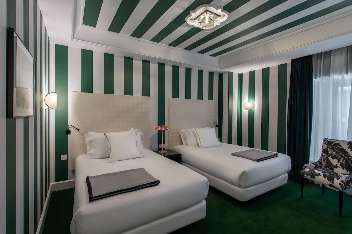 Habitación de hotel con dos camas, paredes verde blanco a rayas, alfombra verde y un sillón estampado.