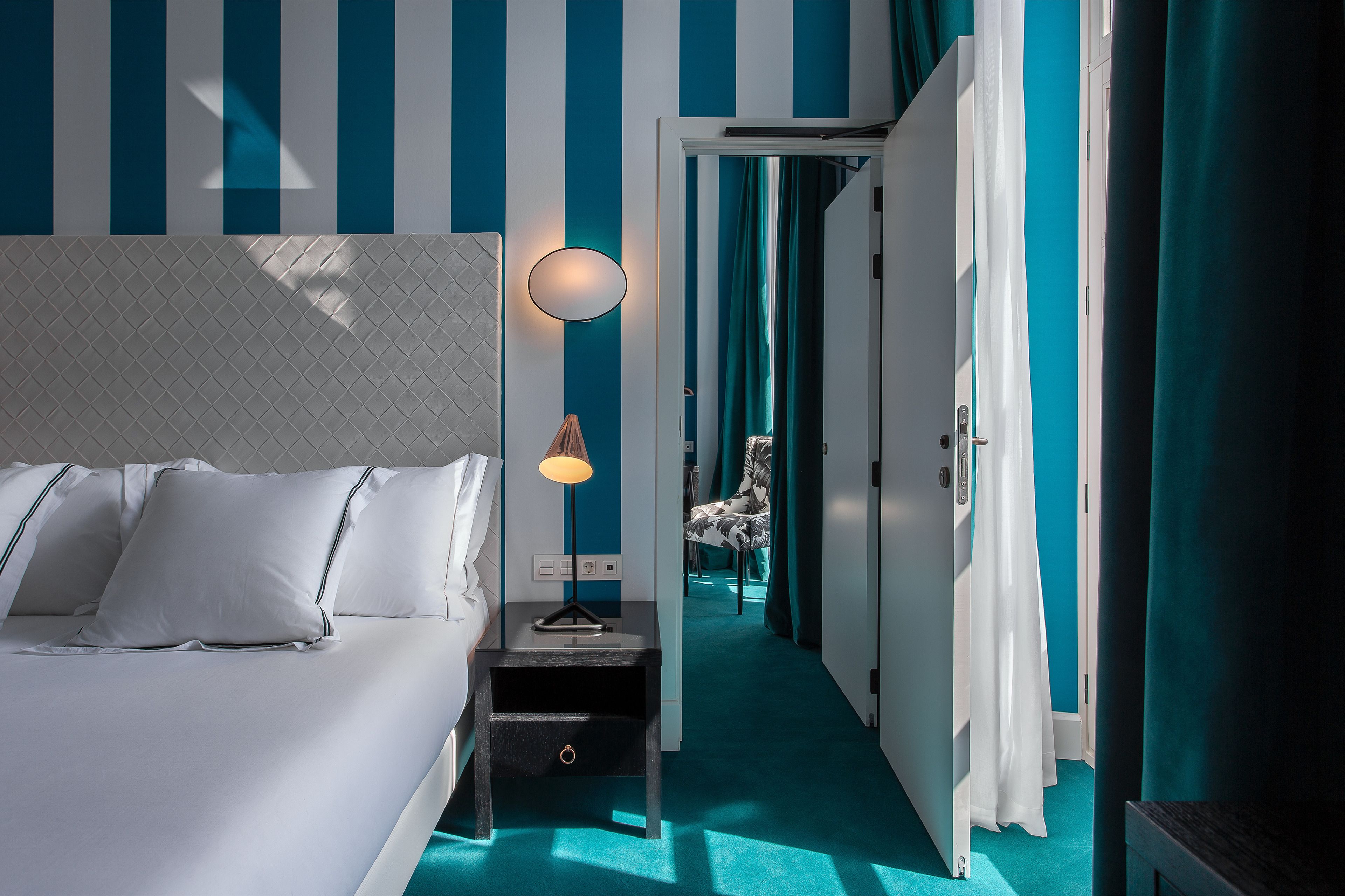 Habitación de hotel con cama, mesita de noche, lámpara, silla y vista a un pasillo con cortinas color turquesa.