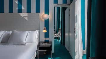 Habitación de hotel con cama, mesita de noche, lámpara, silla y vista a un pasillo con cortinas color turquesa.