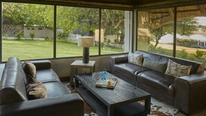 A modern living room with a leather sofa, a coffee table, and a large window offering a view of a garden. - Thamalakane River Lodge (Maun)