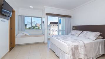 Une chambre d’hôtel moderne dotée d’un grand lit, d’un téléviseur fixé au mur, d’une fenêtre donnant sur la ville et d’un balcon avec vue sur les immeubles.