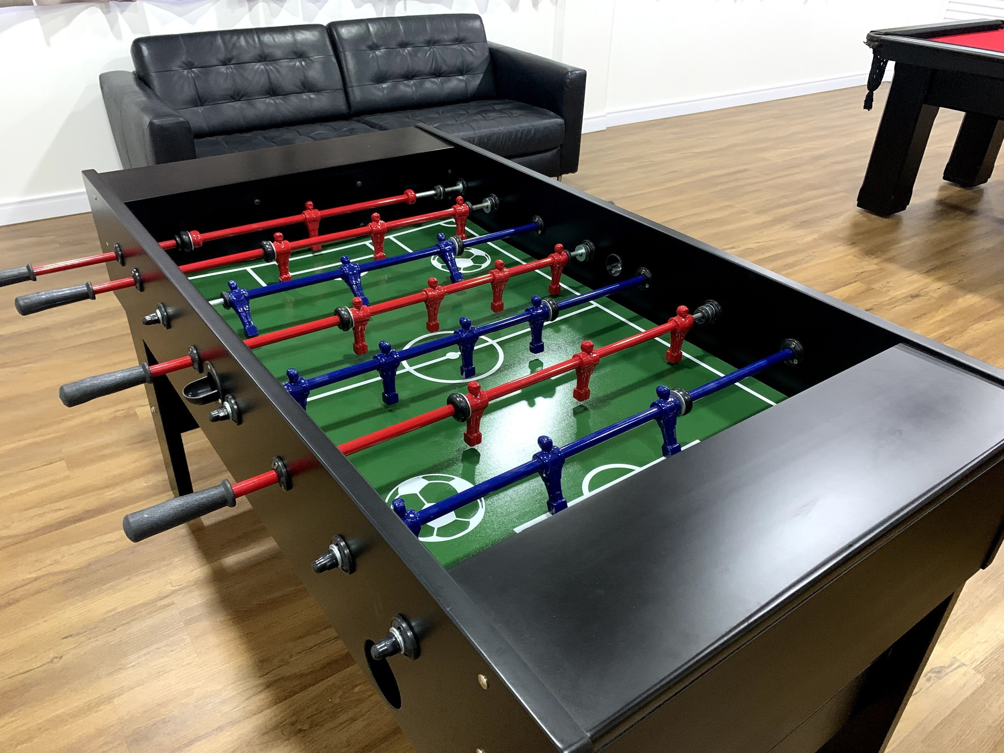 A foosball table in a room with a wooden floor and a black leather sofa.