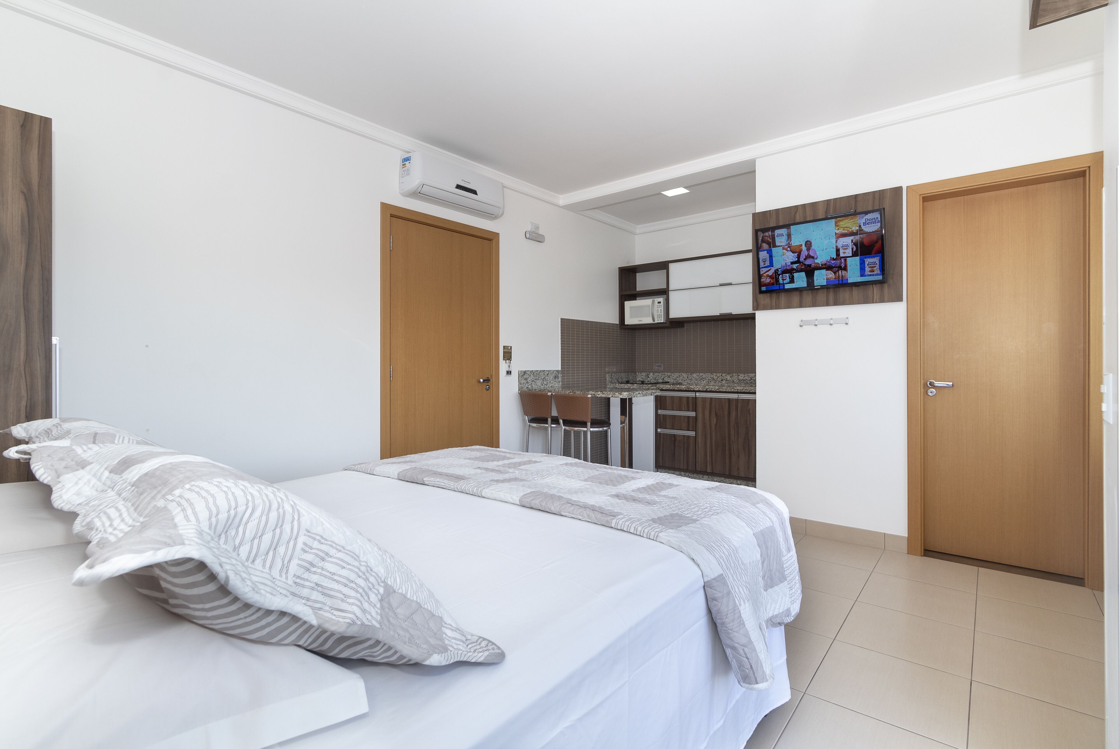 A bedroom with a bed, a TV mounted on the wall, and a kitchenette in the background.