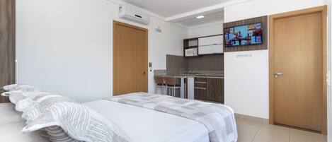 Un dormitorio con cama, televisor montado en la pared y una kitchenette al fondo.
