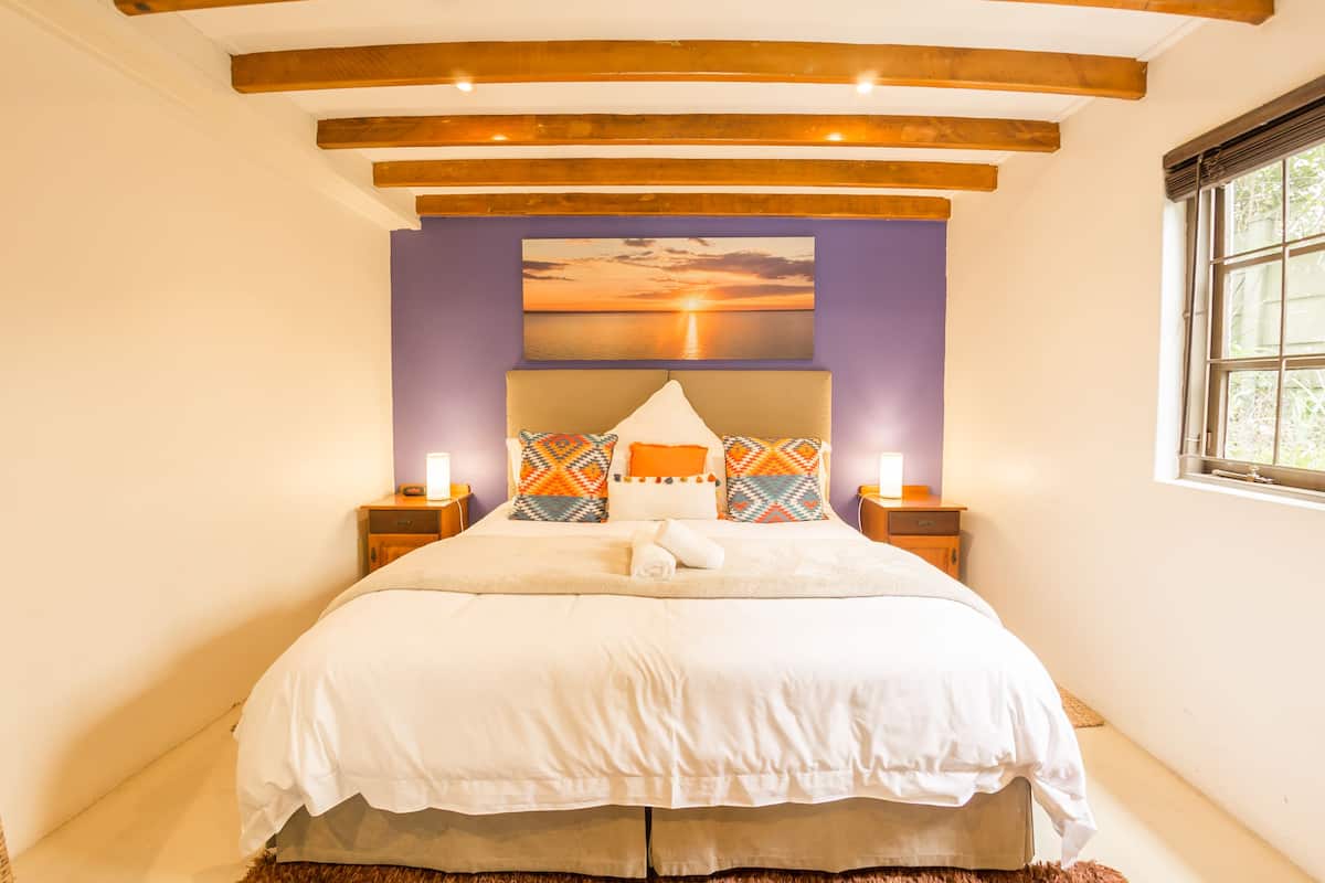 Une chambre avec un grand lit, des poutres en bois au plafond, un tableau représentant un coucher de soleil et deux lampes de chevet.