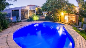 Una piscina con borde de azulejos, rodeada por un jardín y un edificio con antena parabólica.
