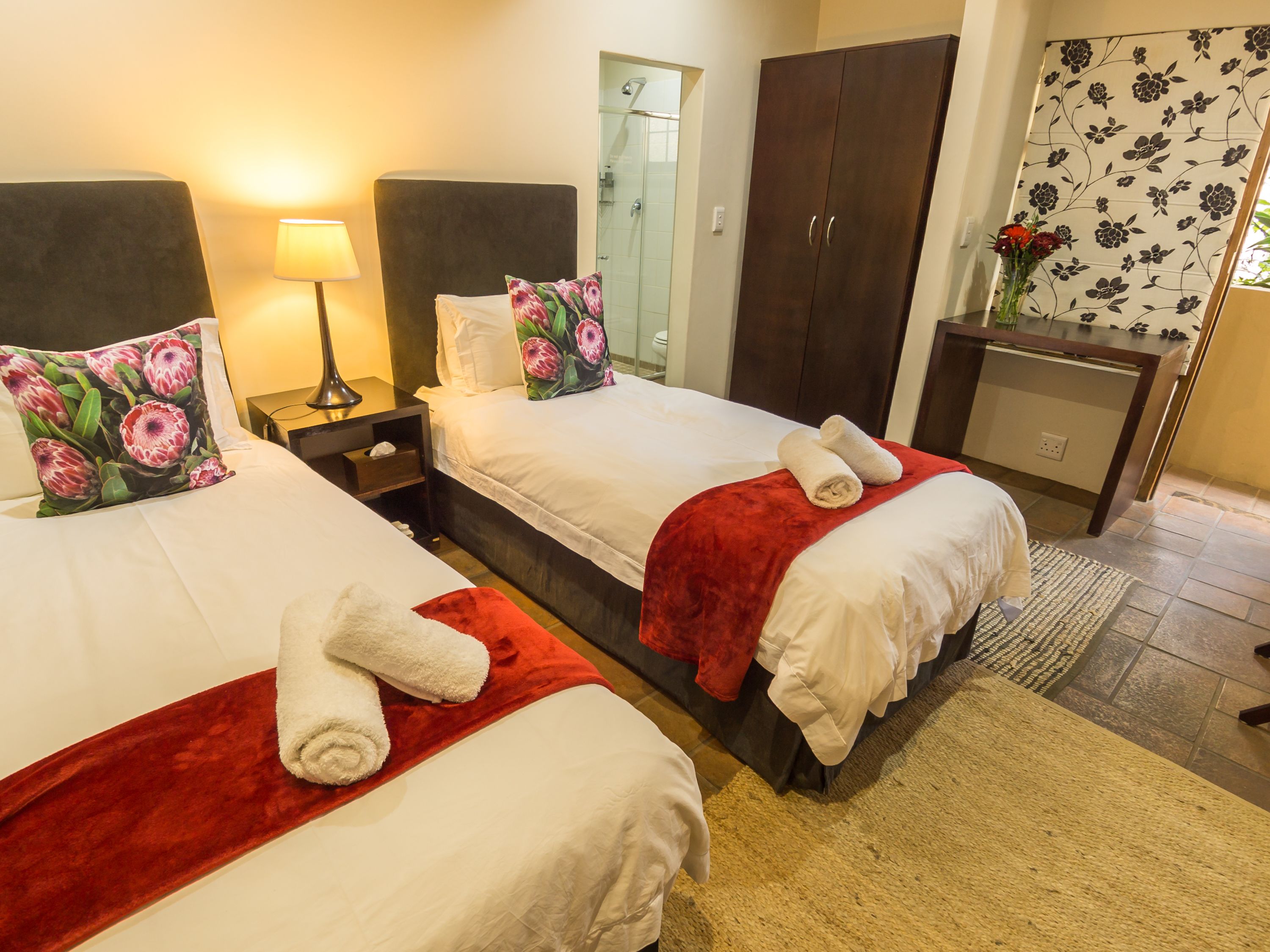 A hotel room with two beds, each with a red blanket and white linens, a nightstand with a lamp, a wooden wardrobe, and a small desk with a vase of flowers.