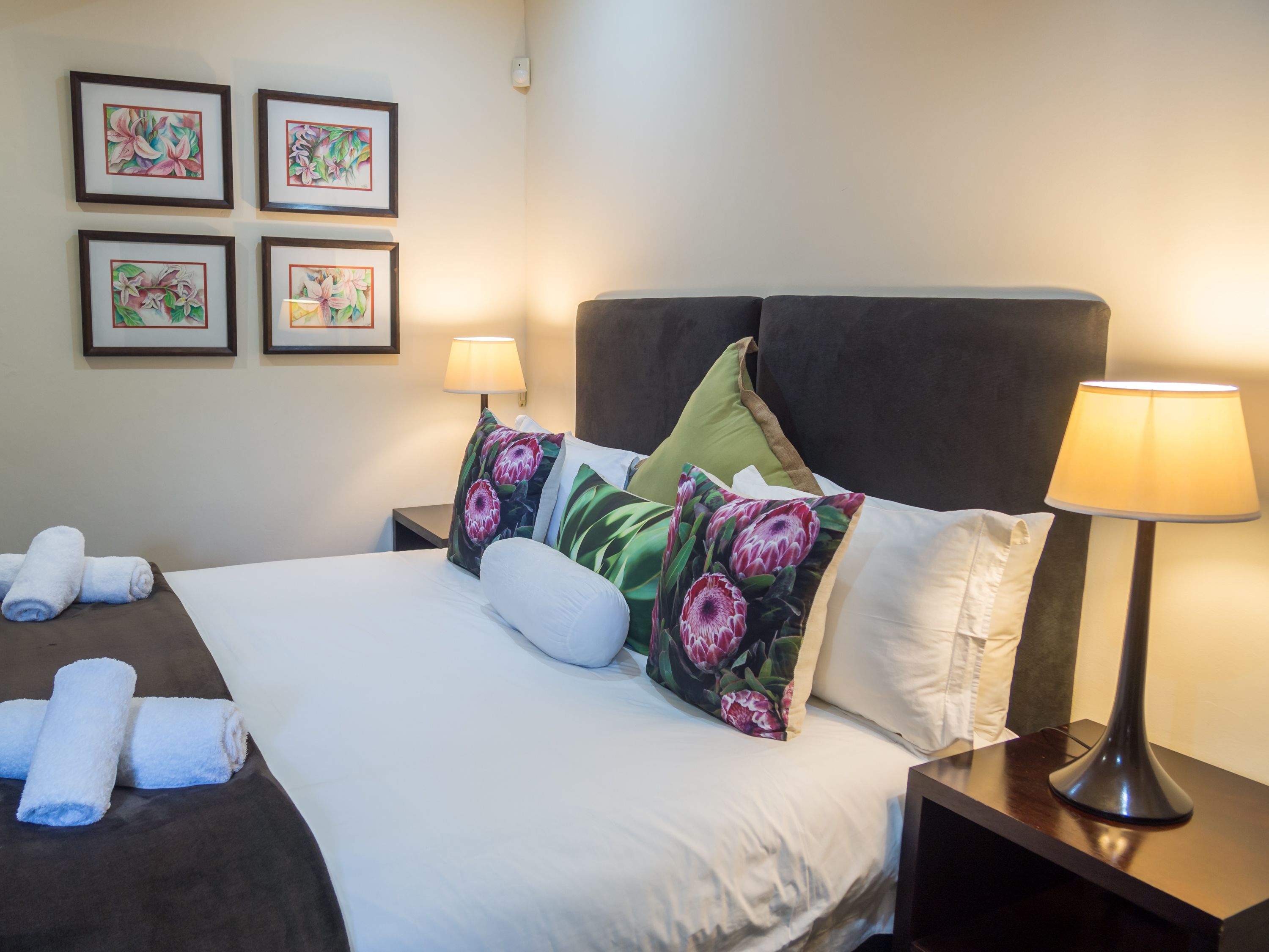 A neatly made bed with floral pillows, a bedside lamp, and a wall with framed floral artwork.