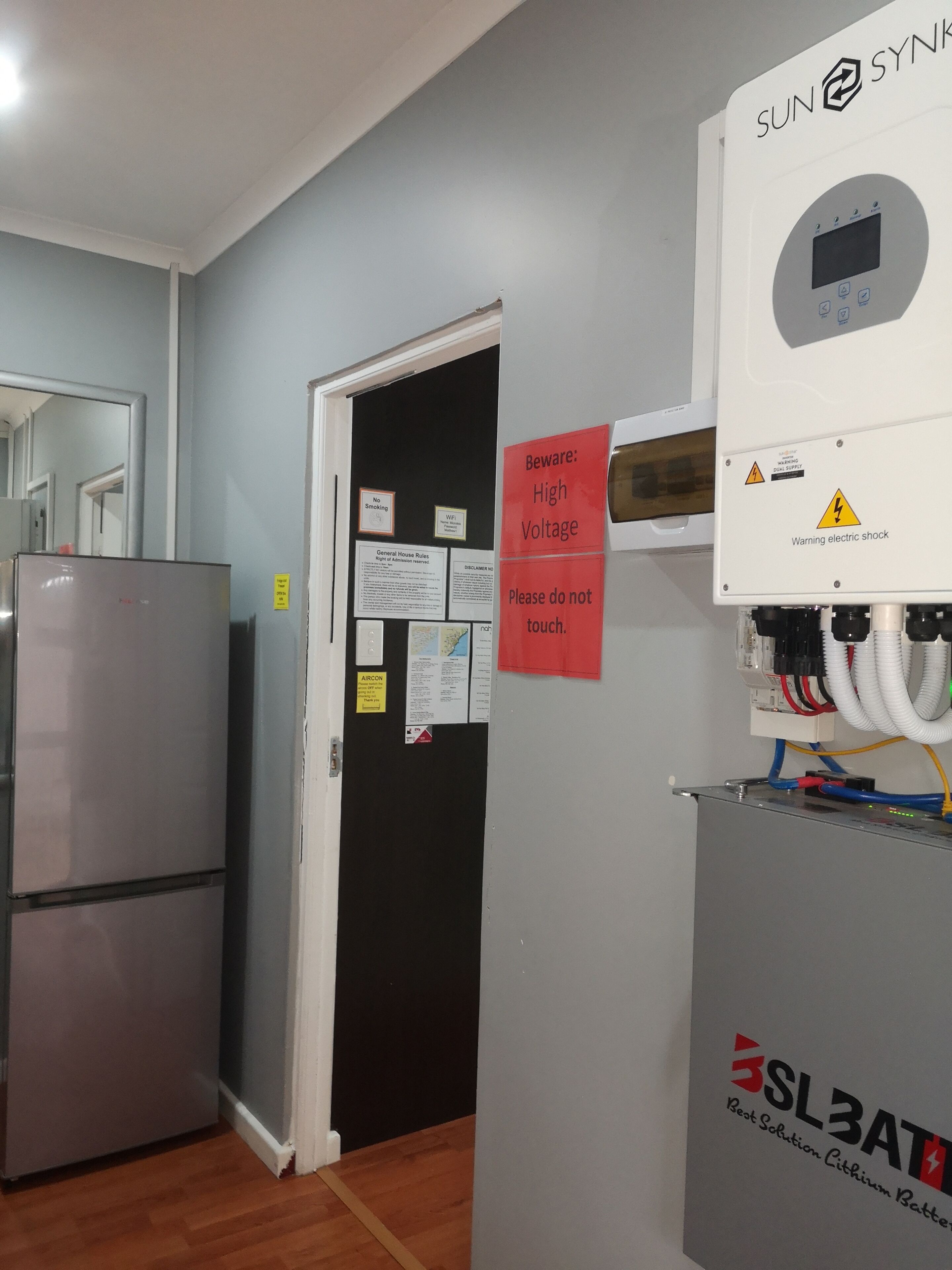 Une pièce équipée d’un réfrigérateur, d’un tableau électrique fixé au mur et d’une porte sur laquelle est apposé un panneau d’avertissement.