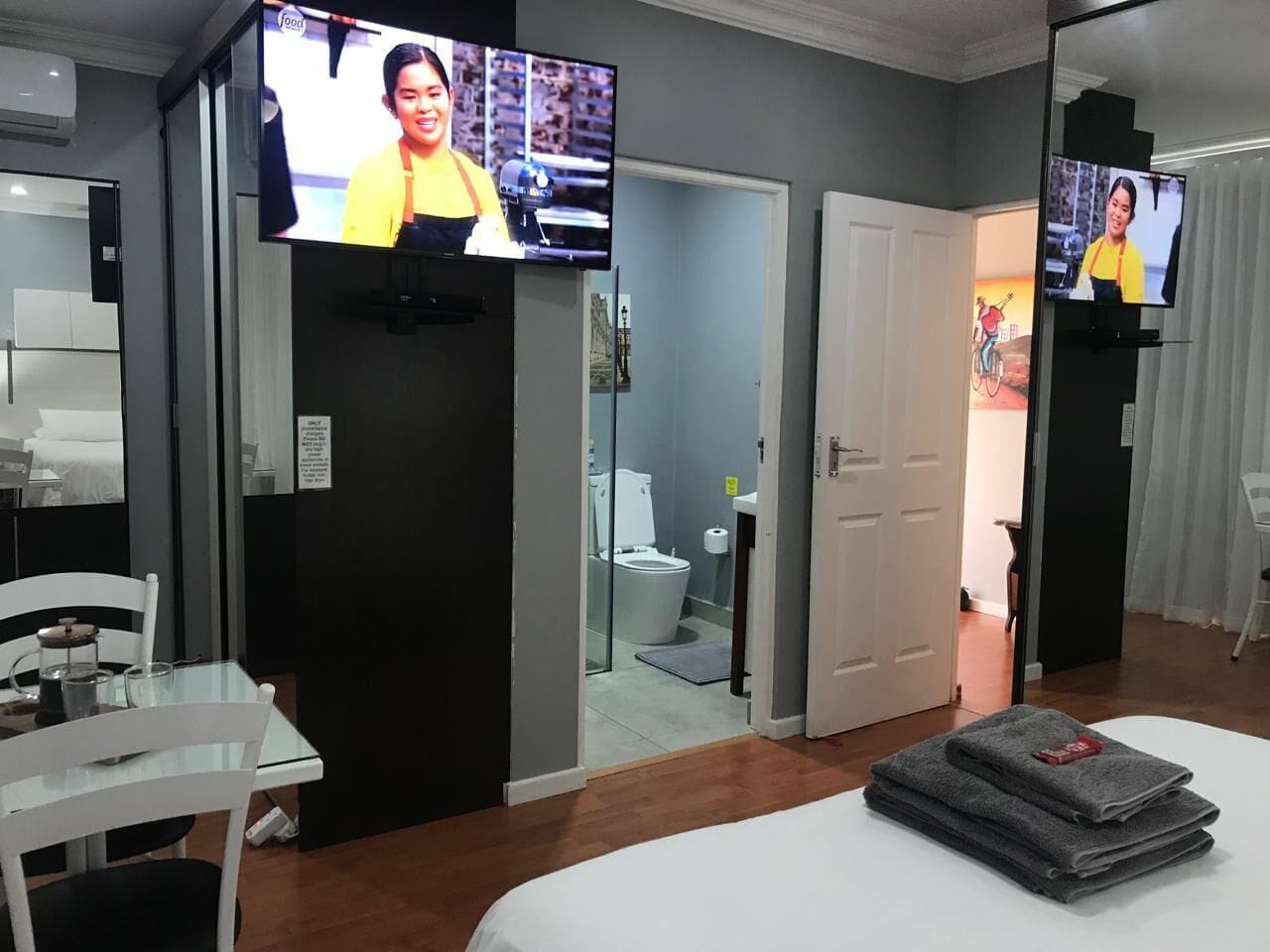 Habitación de hotel con cama, televisor montado en la pared, una mesita con silla y baño visible a través de una puerta abierta.