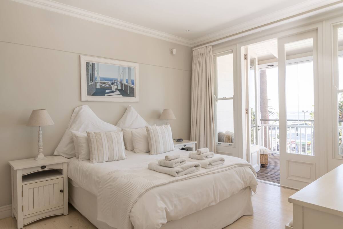 Een hotelkamer met een groot bed, witte lakens, een nachtkastje met een lamp, een lijst met een afbeelding aan de muur, en uitzicht op de omgeving door een deur.