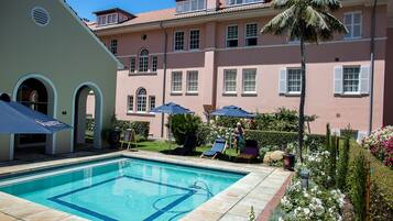 Relax by the rectangular pool surrounded by a tiled deck and lounge chairs, set amidst lush gardens and a classic building.