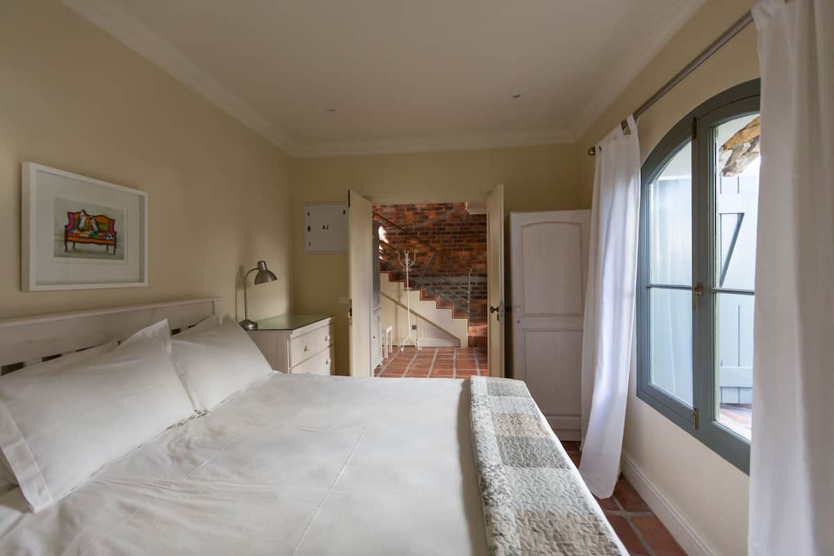 Een slaapkamer met een groot bed, een nachtkastje, een lamp, een ingelijst schilderij aan de muur, en uitzicht op een trap naar een balkon.