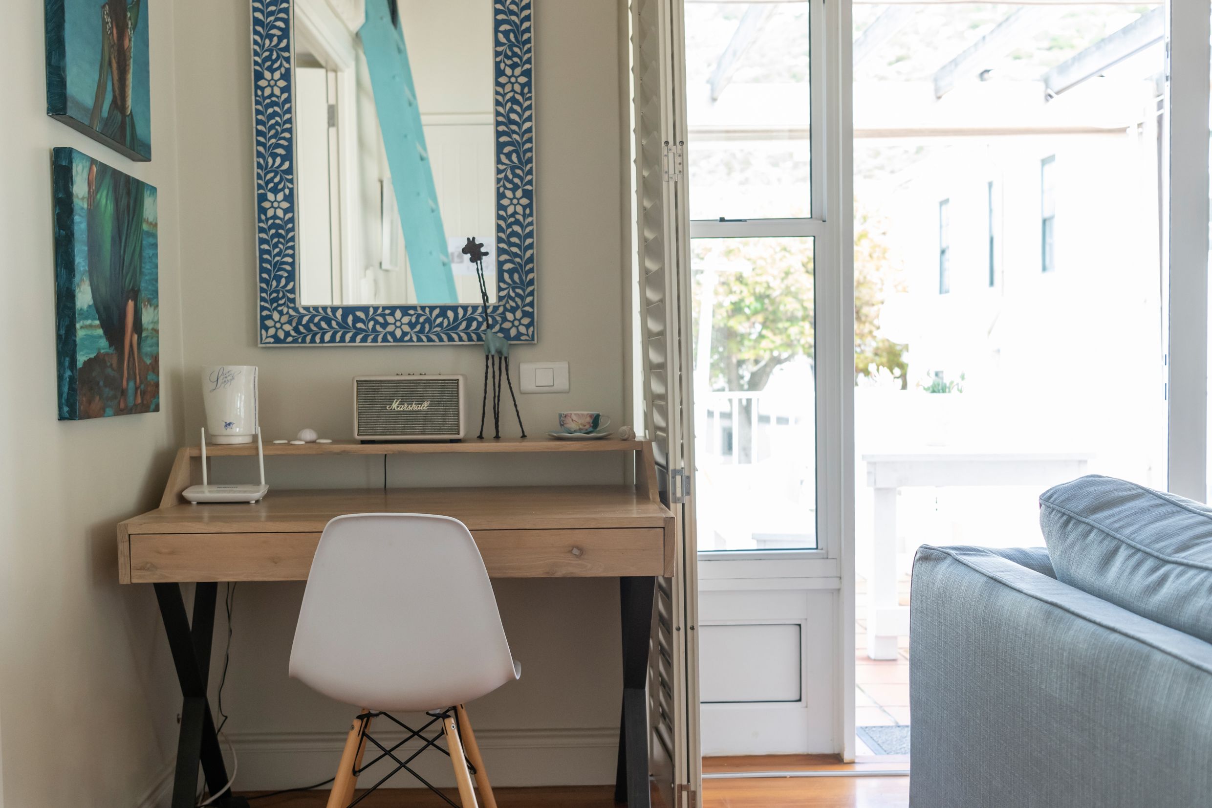 A room with a wooden desk, a white chair, a large mirror with a blue frame, and a view of the outside through a large window.