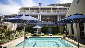 Een zwembad met ligbedden en parasols, een groot huis met balkon, en een bergachtige achtergrond.