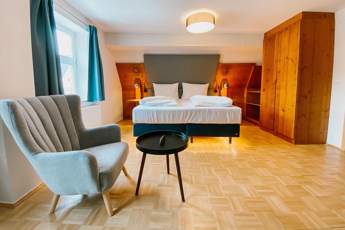 A bedroom with a bed, a chair, a small table, wooden flooring, and a window with curtains.
