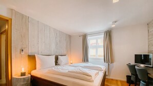 A bedroom with a wooden headboard, a bed with white linens, a bedside table with a lamp, a desk with a chair, and a window with curtains.