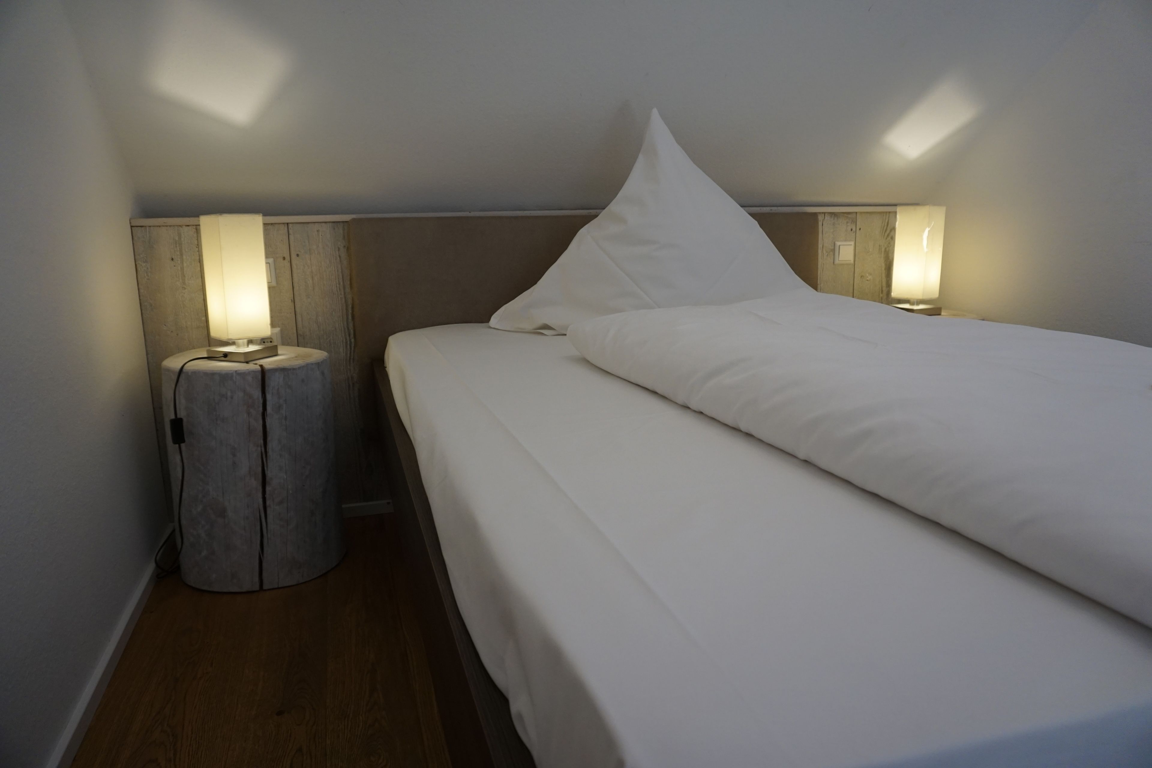 A small, dimly lit room with a single bed, a nightstand, and a wall-mounted lamp.
