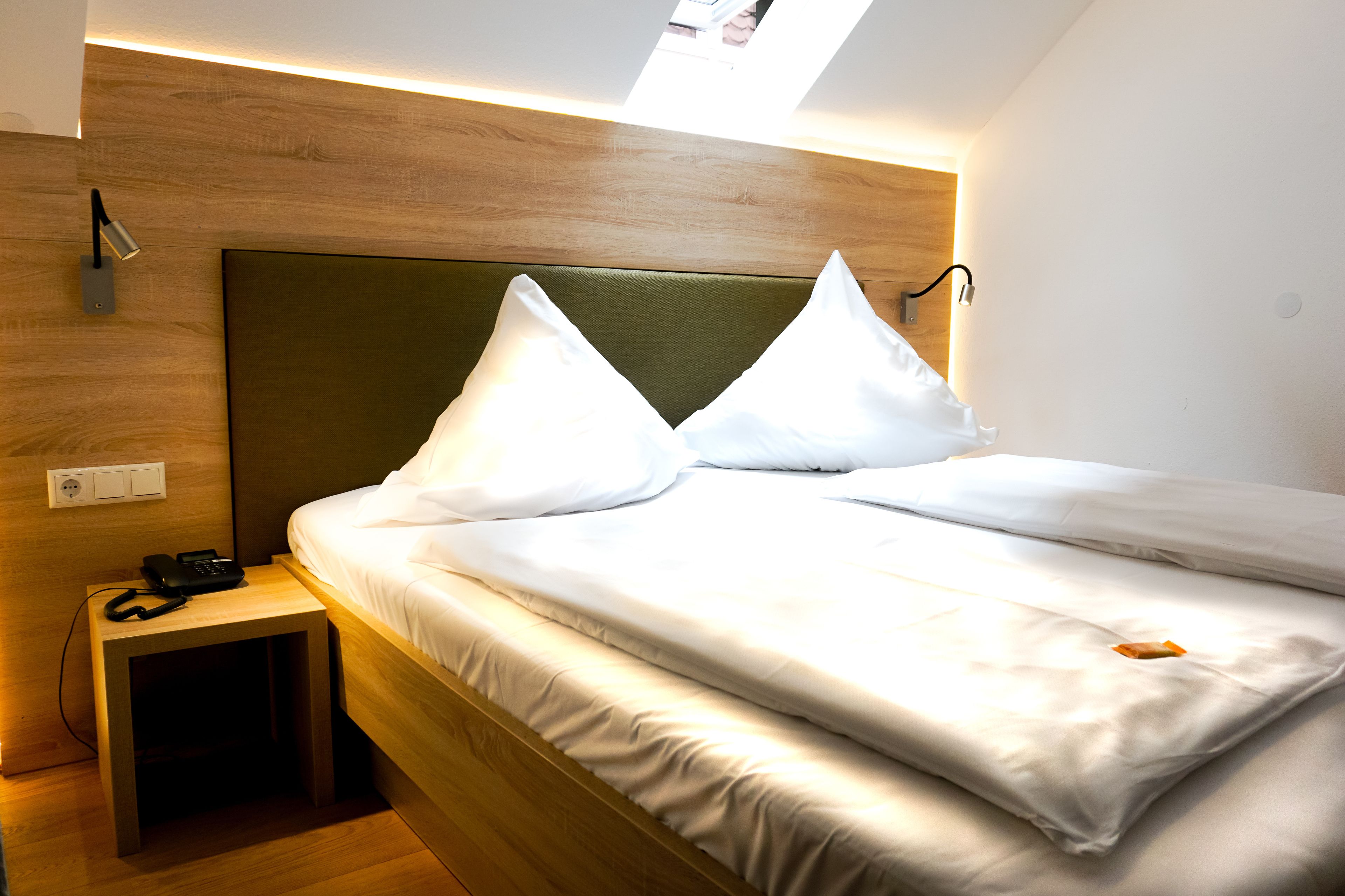 Un lit bien fait, avec du linge de lit blanc et des oreillers, une table de chevet avec un téléphone et un luminaire fixé au mur.
