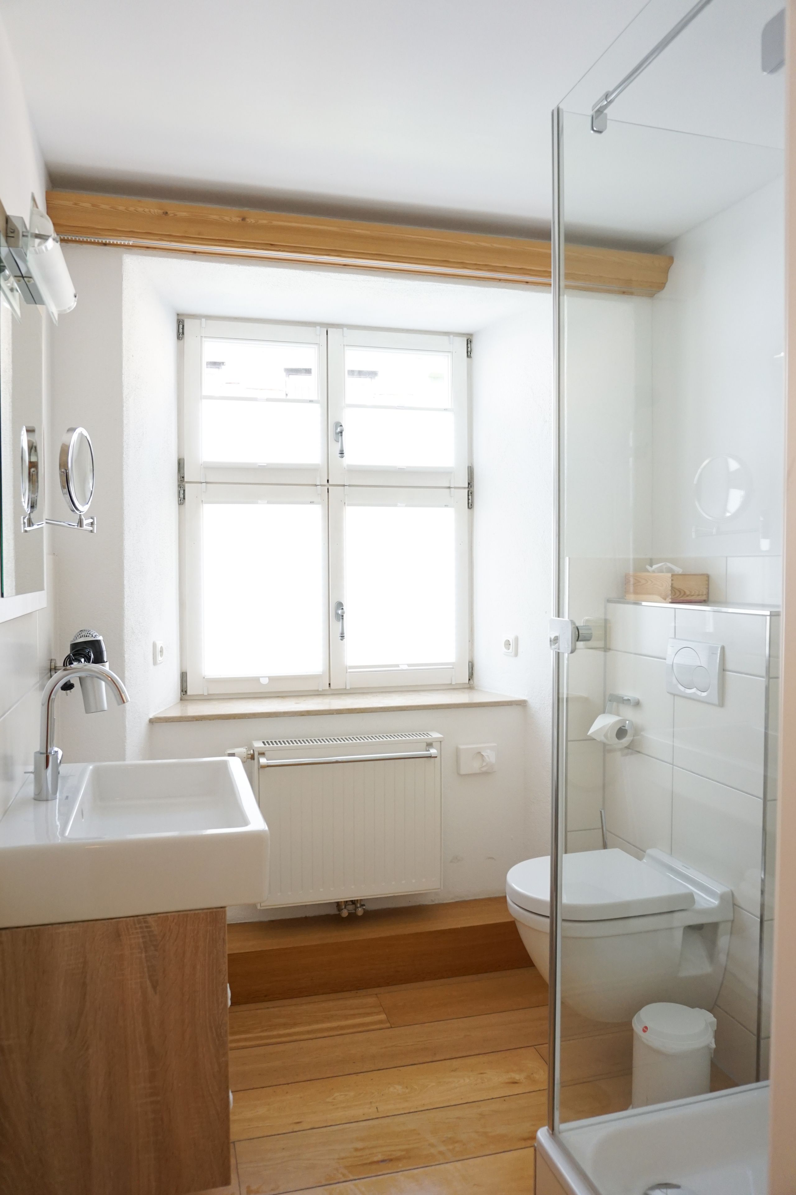 A modern bathroom with a wooden floor, white fixtures, a window, and a glass shower enclosure.