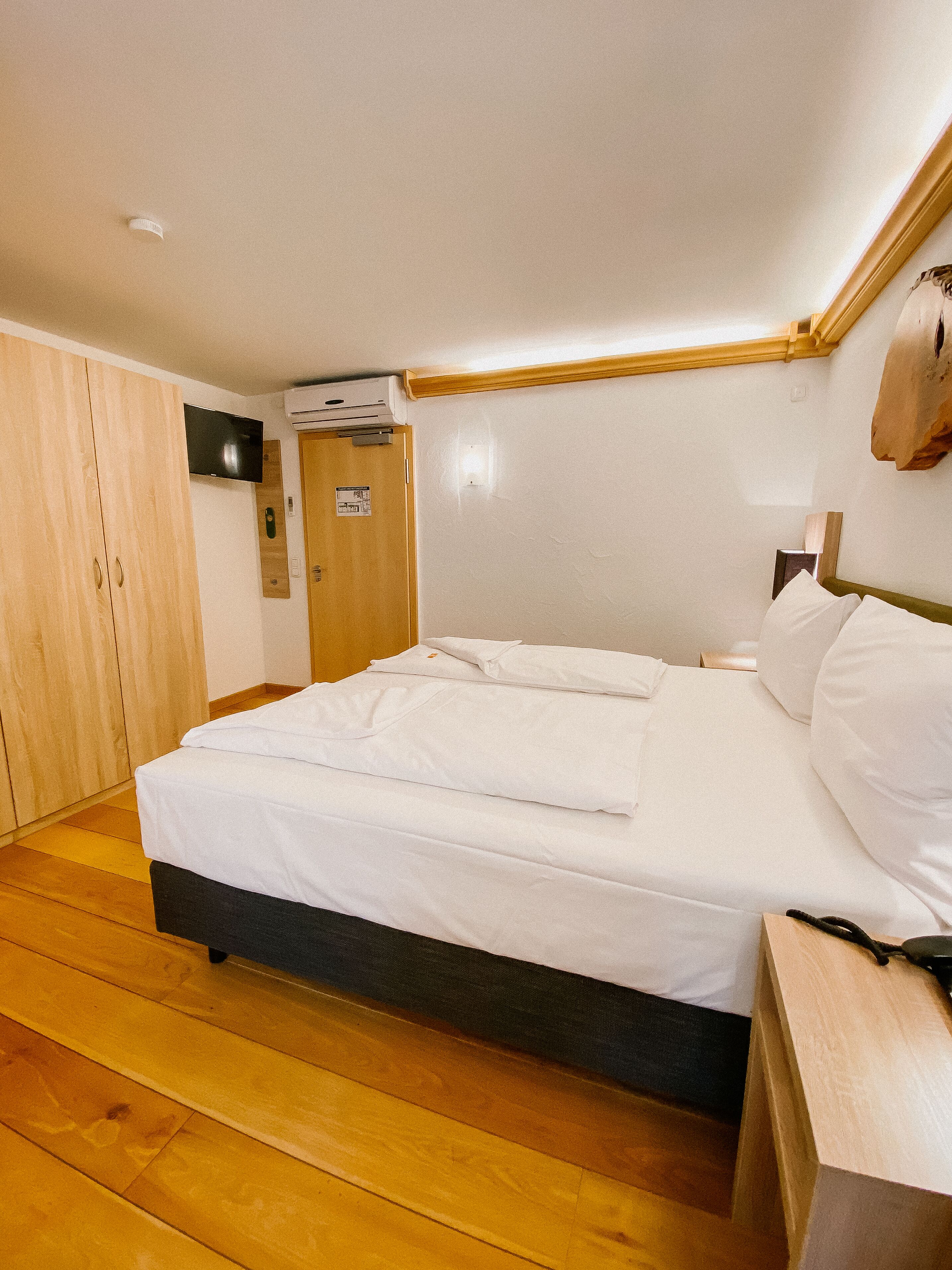 Un dormitorio con suelo de madera, una cama con ropa de cama blanca, un armario de madera, un televisor de pantalla plana y un equipo de aire acondicionado.