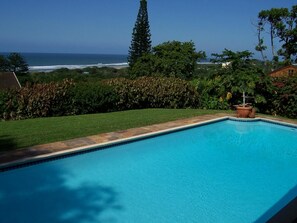 A clear blue swimming pool with a view of the ocean and a well-maintained garden. - Birdcage B and B (Kingsburgh)