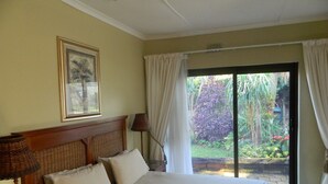A bedroom with a large bed, a headboard, a framed picture, a window with curtains, and a view of a garden. - Birdcage B and B (Kingsburgh)