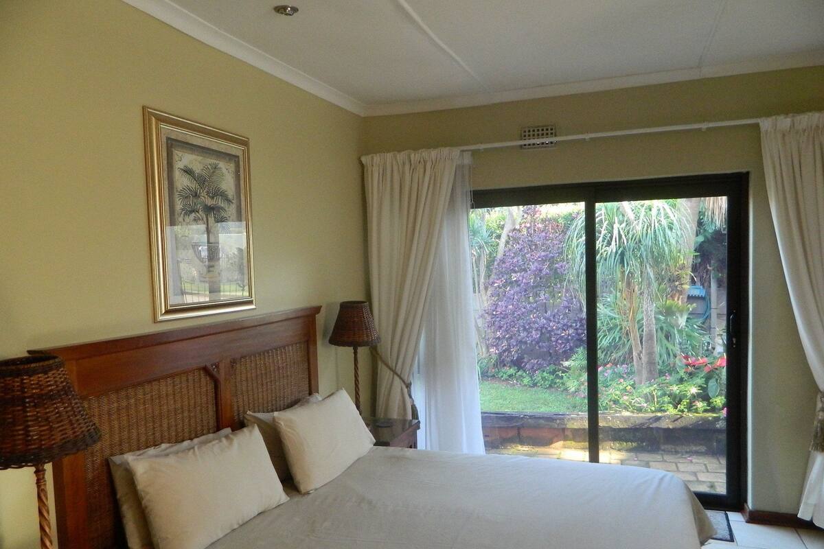 A bedroom with a large bed, a headboard, a framed picture, a window with curtains, and a view of a garden.